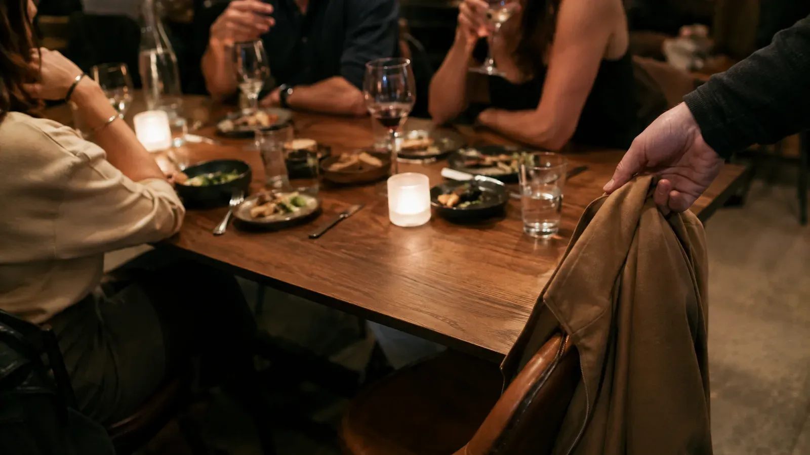 A restaurant table with several people talking and one empty chair, a coat being lifted from its back